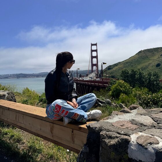 Katalina sitting cross-legged on a wooden beam, looking toward the Golden Gate Bridge